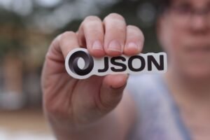 jQuery Certification Level 1 - A close-up of a hand holding a JSON logo sticker outdoors, blurred background.