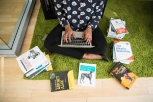Professional Neuro Linguistic Programming Certificate - Woman sitting on green rug working on laptop, surrounded by technology books in a modern room.