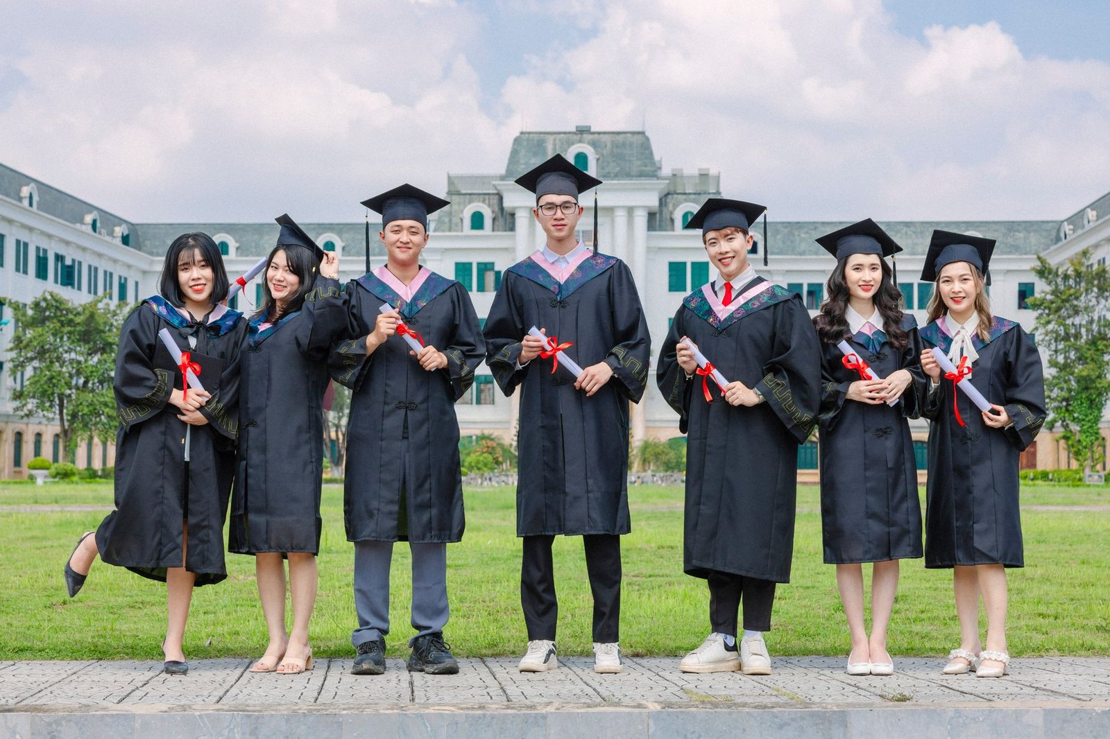 Recruitment Diploma - Group of diverse young graduates in gowns holding diplomas outdoors in front of a university building.