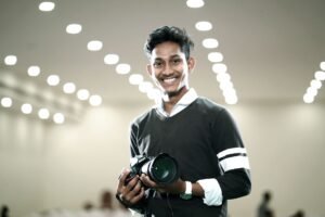 Confidence On Camera Certification - Confident young man holding camera against a backdrop of modern lights, smiling warmly.