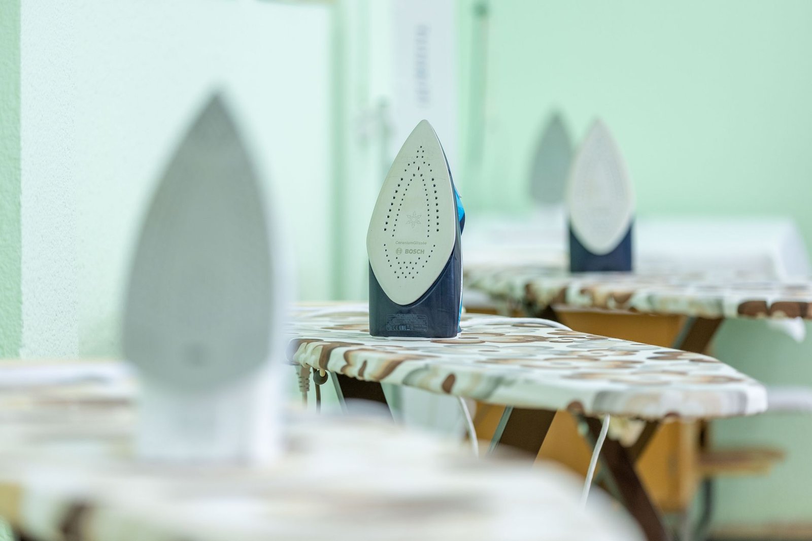 Start Your Own Ironing Business Diploma - Close-up of modern irons placed on ironing boards with camouflage pattern covers in a bright indoor setting.