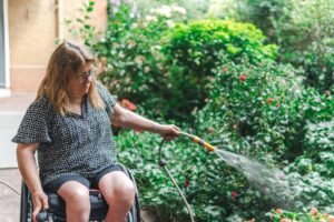 Introduction to Independent Living Certification - A woman in a wheelchair tending to her garden, watering plants with a hose.
