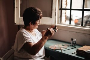 Stroke Awareness Certification - Senior woman engaged in painting by a window, with art supplies on the desk, creating art in natural light.
