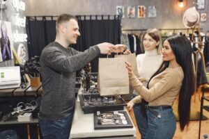 Customer Service in Fashion Shops Certification - Side view of cheerful male shop assistant standing at counter desk and giving to positive female customers paper bags with purchases in trendy shop
