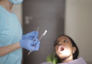 Oral Health in Care Certification - Unrecognizable crop dentist in medical mask and latex gloves wearing blue uniform holding instruments while treating teeth of female patient in modern clinic