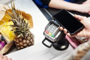 Mobile and Portable Device Security Certification - Close-up of a customer using a smartphone for contactless payment at a retail checkout with a pineapple on the counter.
