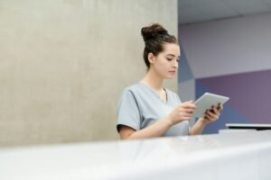 Medical Receptionist Administration Certification - A receptionist in scrubs using a tablet at an office reception desk.