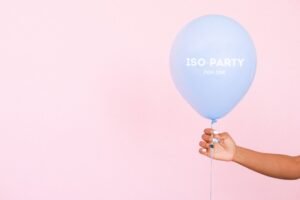 Understanding ISO 27001 - A hand holding a blue balloon with 'ISO-PARTY FOR ONE' text on a light pink background.