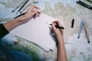 Travel Writing Certification - Hands writing on paper over a detailed world map with various pens.