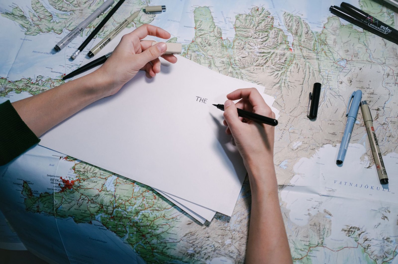 Travel Writing Certification - Hands writing on paper over a detailed world map with various pens.