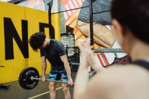 Commitment to Consistency Certification - A young man performs a deadlift in a vibrant gym, watched by a supporter.