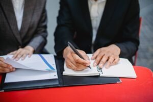 Minute Taking Certification - Two professionals in suits writing notes and reviewing documents at an outdoor meeting table.