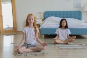 Mental Wellbeing in Children and Young People Certification - Full body of concentrated diverse girls meditating on carpets in bright cozy room next to mirror and big bad in daylight
