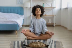 Introduction to Mindfulness for Children Certificate - Full body of African American girl with curly hair meditating with eyes closed and legs crossed in bright cozy bedroom