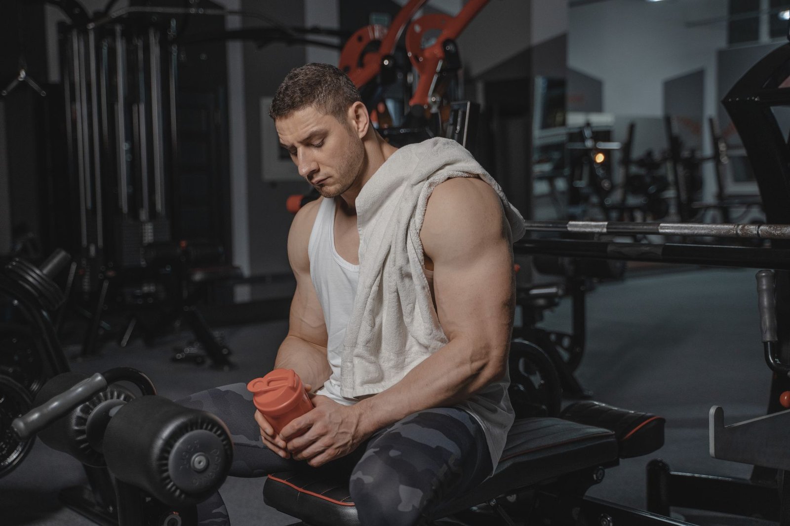 Introduction to Sports Nutrition Certification - Muscular man sits in gym holding protein shaker after a workout, towel on shoulder.