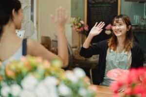 Meeting and Greeting Customers Certification - Positive Asian woman greeting client waving hand while working as florist in flower shop