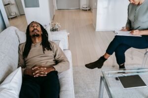 Introduction to REBT: Rational Emotive Behaviour Therapy Certification - From above of African American male looking up while lying on couch near crop female therapist writing notes on clipboard during session