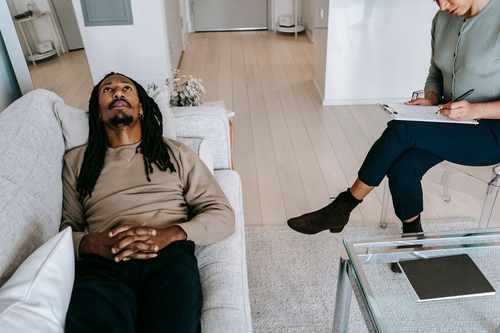 Introduction to REBT: Rational Emotive Behaviour Therapy Certification - From above of African American male looking up while lying on couch near crop female therapist writing notes on clipboard during session