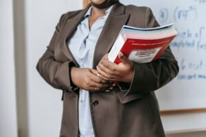 The Key Stages of the National Curriculum Certification - Crop faceless African American female teacher in formal clothes standing near whiteboard with textbooks in hand during lesson in classroom