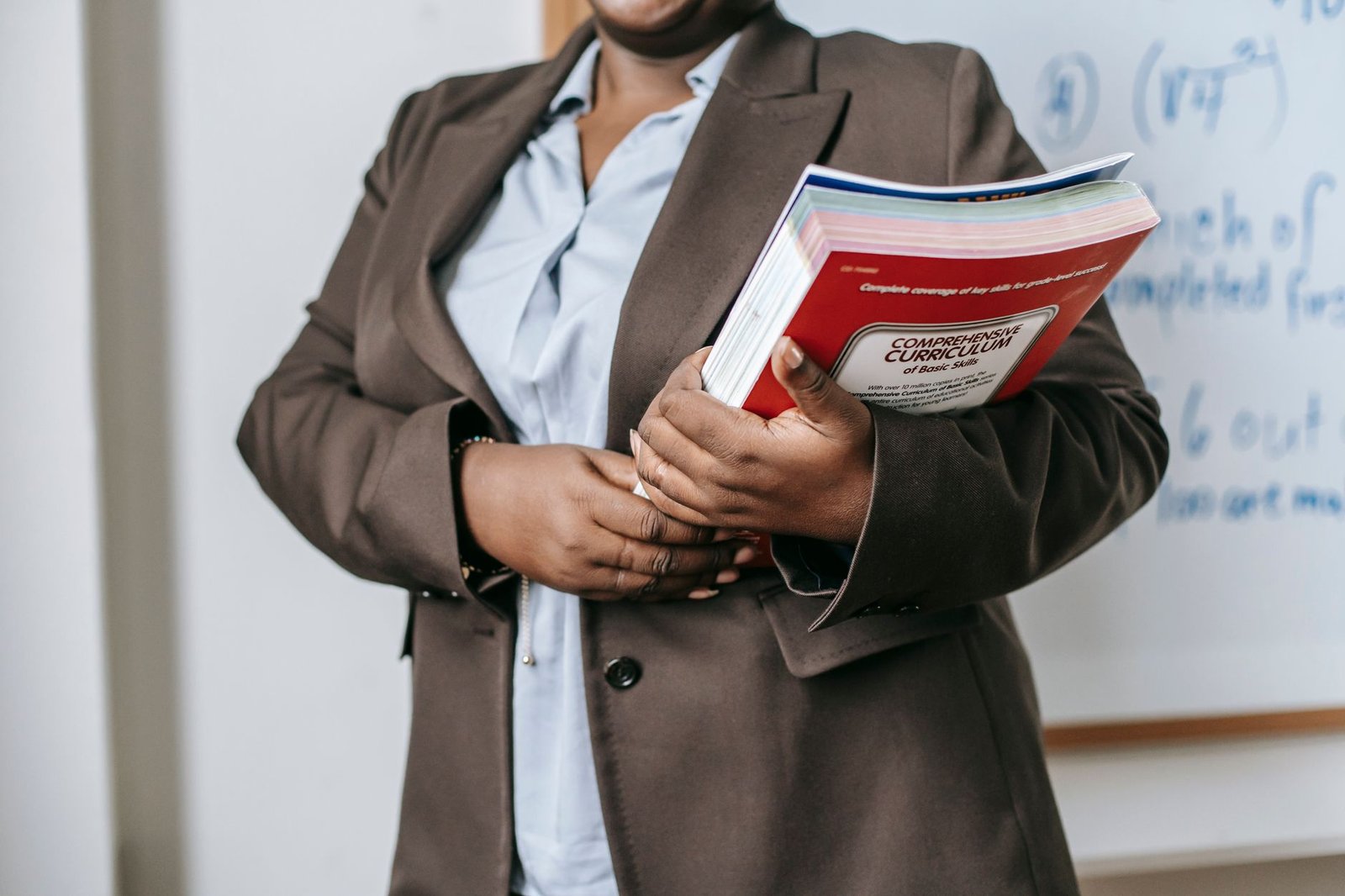 The Key Stages of the National Curriculum Certification - Crop faceless African American female teacher in formal clothes standing near whiteboard with textbooks in hand during lesson in classroom