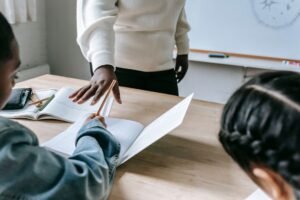 Becoming a Higher Level Teaching Assistant Certification - Crop faceless woman standing near desk while schoolchildren writing test with pencils and copybooks in classroom