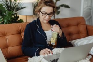 Alcohol at Work Awareness Certification - Woman in pajamas holding a bottle of alcohol while working on a laptop, sitting on a sofa at home.