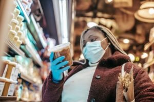 Food Safety Level 2 Certification - Low angle of African American female in mask and latex gloves considering food while shopping in supermarket during pandemic of coronavirus