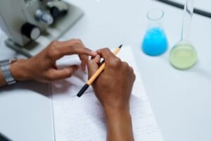 Researching Certification - Close-up of a scientist taking notes in a laboratory setting with flasks and a microscope.