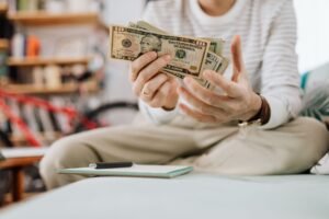 How to Manage Your Money Certificate - Man wearing casual clothes counts dollar bills while sitting on a sofa indoors.