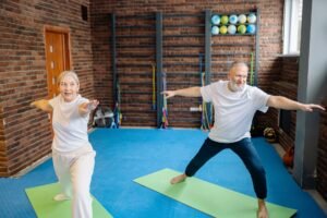 Preventing Falls in Older People Certification - Elderly couple doing yoga poses on mats in a gym, promoting active lifestyle.