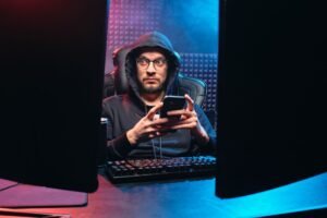 Mobile Phishing Awareness - A man in a hoodie using a smartphone, surrounded by tech gear in a dimly lit room.