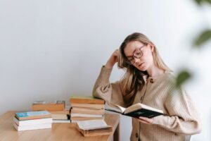Learn Speed Reading Certification - Thoughtful female student in sweater and eyeglasses standing and reading book in light room