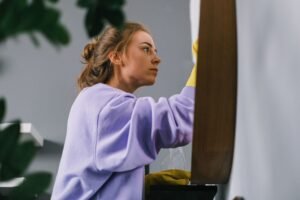 Cleaning Certification - Side view of young focused female in latex gloves standing and cleaning mirror in light room in daytime