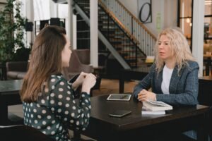 Questioning and Listening Techniques Certification - Businesswomen sitting at table with book and devices and having conversation about project in modern workplace