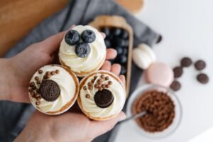 Cupcake Academy: Introduction to Running a Baking Business - Top view of crop anonymous person showing delicious cupcakes with blueberries and chocolate sweets against blurred background of various dessert