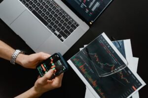 Digital Marketing Certification Level 2 - Hands using smartphone beside laptop with stock charts, showcasing digital trading.