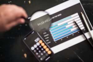Sales Tools and Technology Certification - Close-up of a magnifying glass focusing on a sales volume chart next to a smartphone calculator app.