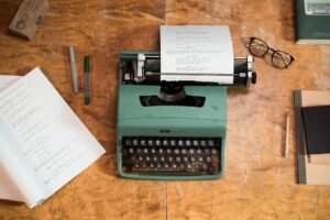 Script Writing Certification - A vintage green typewriter with scripts surrounded by glasses and pens on a wooden table.