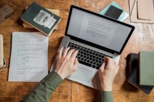 Copywriting Masterclass Certification - High-angle view of hands typing on a laptop surrounded by books and papers.