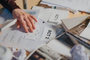 The Freedom of Information Act Certification - A cluttered desk with scripts, notes, and a person's hand organizing screenplay drafts.