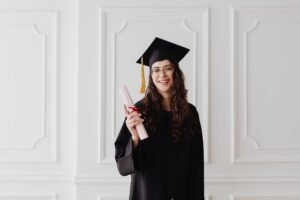 The Complete Menopause Toolkit Certification - Smiling young woman in graduation gown and cap holding a diploma indoors.