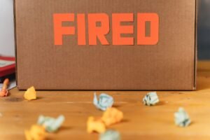Termination of Employment Certification - Close-up of a cardboard box labeled FIRED on a wooden table with crumpled papers.