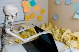 How to Stop Procrastinating Certification - A skeletal figure works at a desk with a laptop and scattered papers, symbolizing burnout.