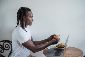 Eat to Gain Certificate - Man enjoys breakfast sandwich while working on laptop at home.