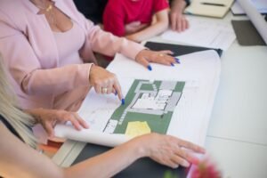 Master Planning Certification - A real estate agent in a pink blazer shows floor plans to clients, discussing property details.