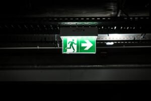 Martyn’s Law and Emergency Preparedness Certification - Bright green emergency exit sign with arrow in a dark industrial setting, indicating evacuation route.