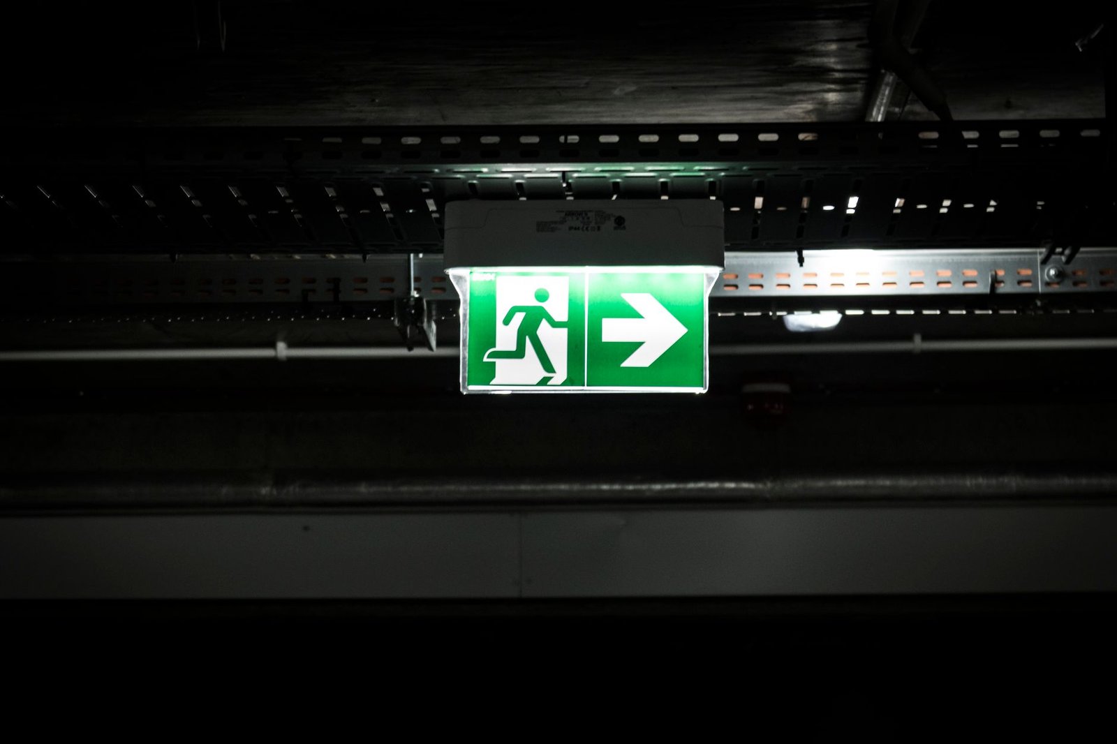 Martyn’s Law and Emergency Preparedness Certification - Bright green emergency exit sign with arrow in a dark industrial setting, indicating evacuation route.
