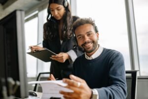 Helpdesk Support Analyst Certification - Friendly call center agent wearing a headset, talking and smiling at his desk with a colleague.