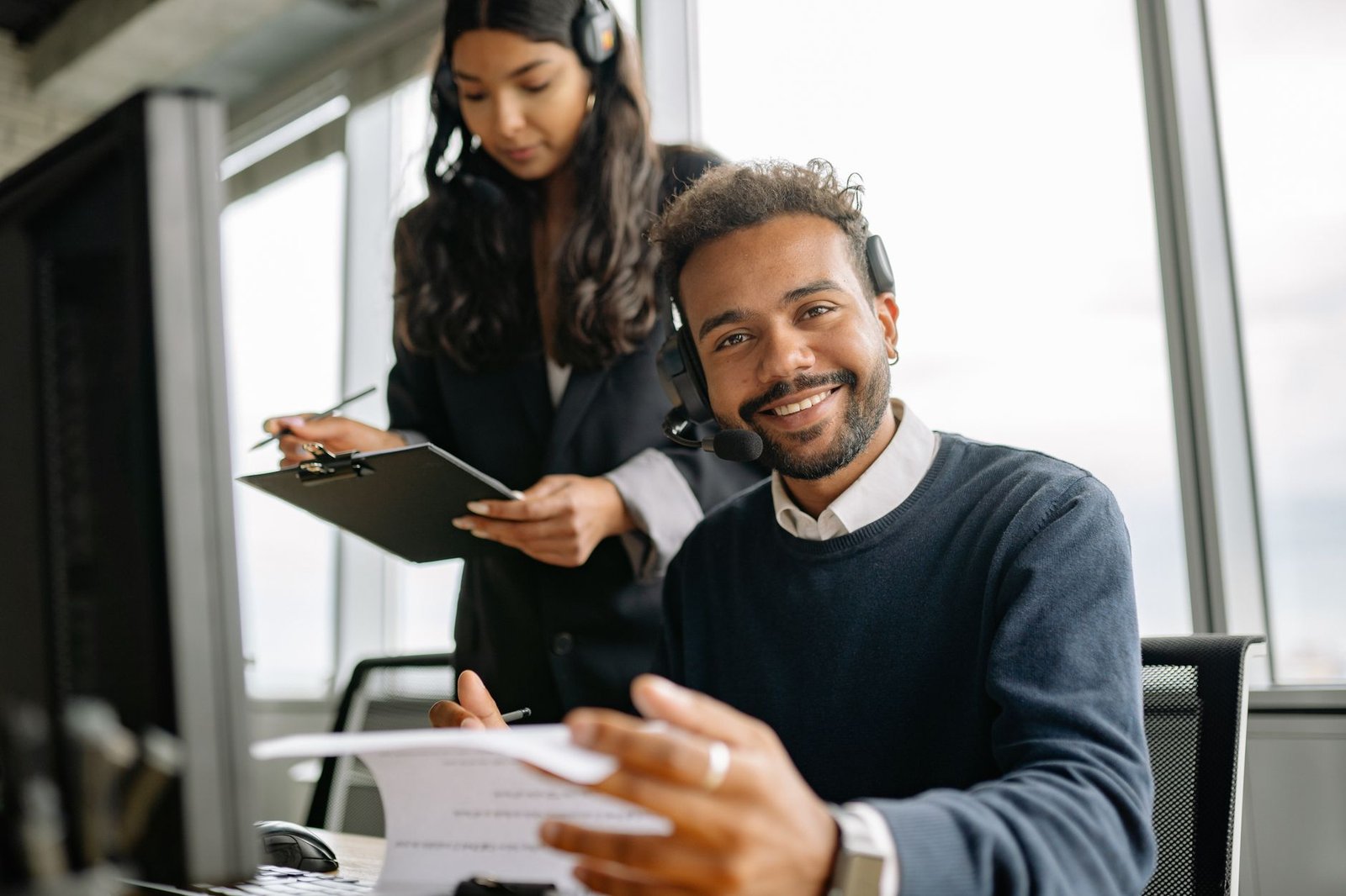Helpdesk Support Analyst Certification - Friendly call center agent wearing a headset, talking and smiling at his desk with a colleague.