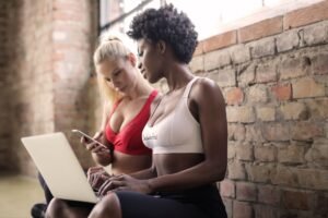 Marketing Your Fitness Business Certification - Two women in sportswear using a laptop and smartphone against a brick wall, showcasing fitness and collaboration.